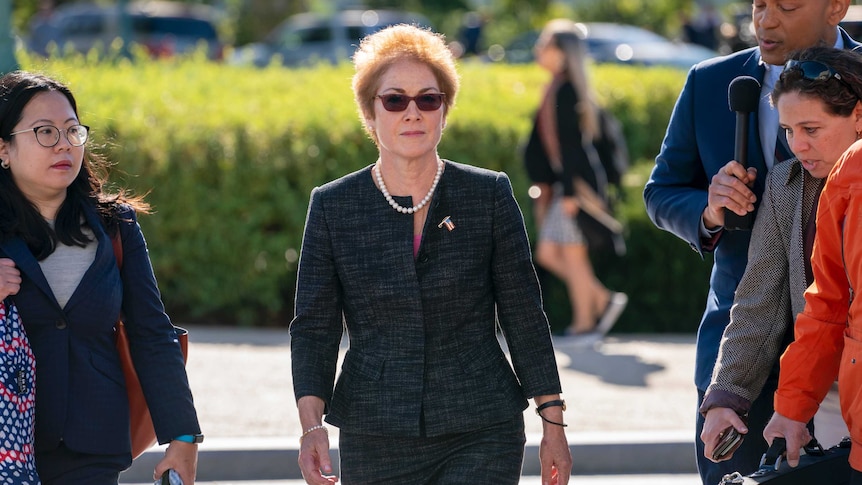Marie Yovanovitch walks towards the camera with  stern expression. She wears sunglasses, a charcoal suit and a US flag brooch.
