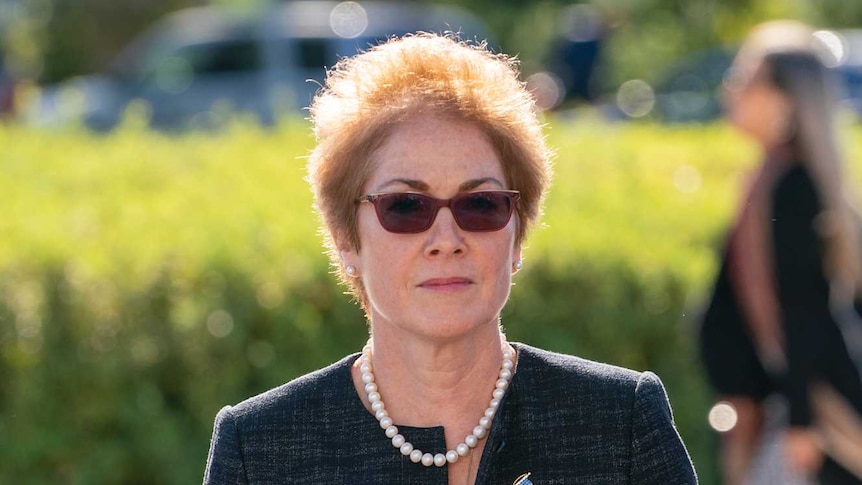 Marie Yovanovitch walks towards the camera with  stern expression. She wears sunglasses, a charcoal suit and a US flag brooch.