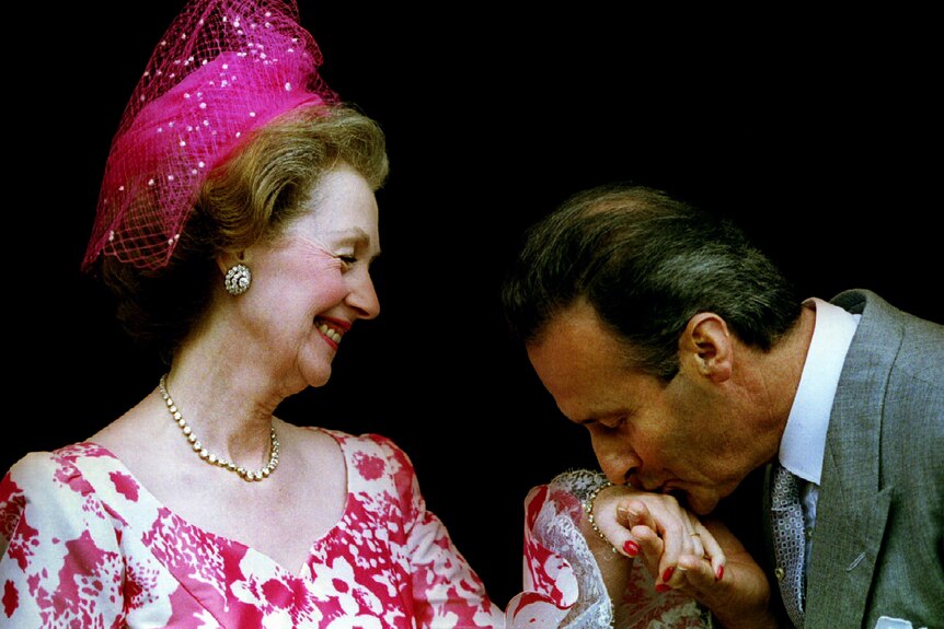 A woman in a pink hat smiles as a man kisses her hand 