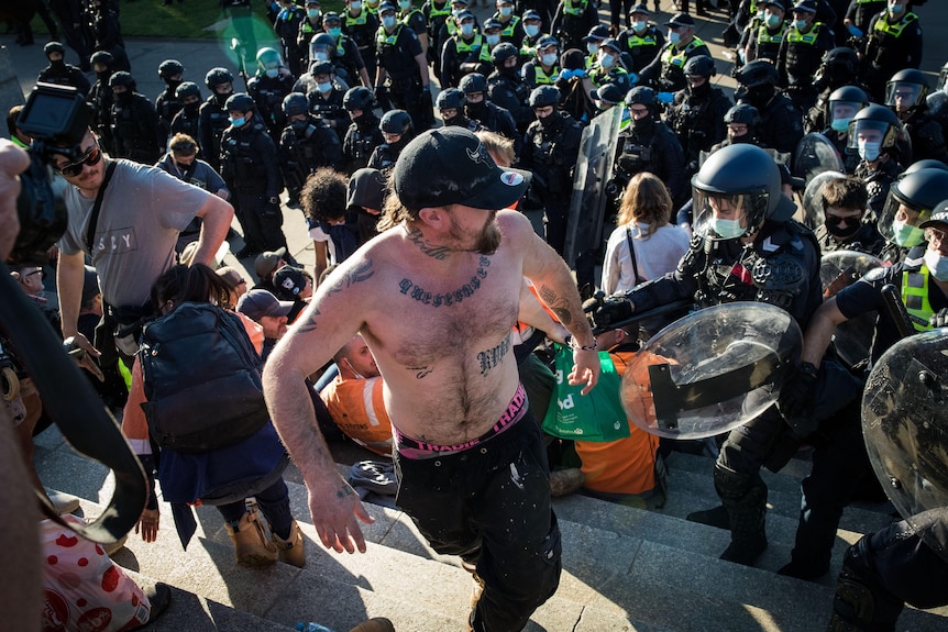 A shirtless tattooed man turns around to face police as they clash with other protestors.