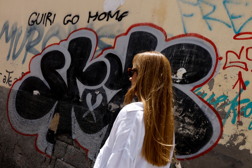 A woman walks past graffiti on the wall in Spain that says "Guiri go home" which translates to "tourist go home".