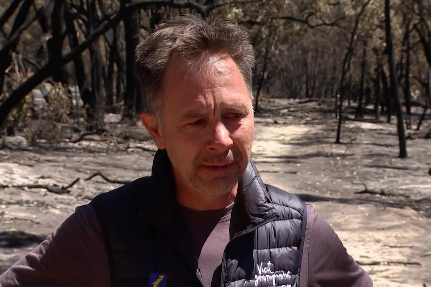 Marc Sleeman wears a dark sleeveless puffer vest and black long sleeve shirt and looks upset while standing in burnt bushland.