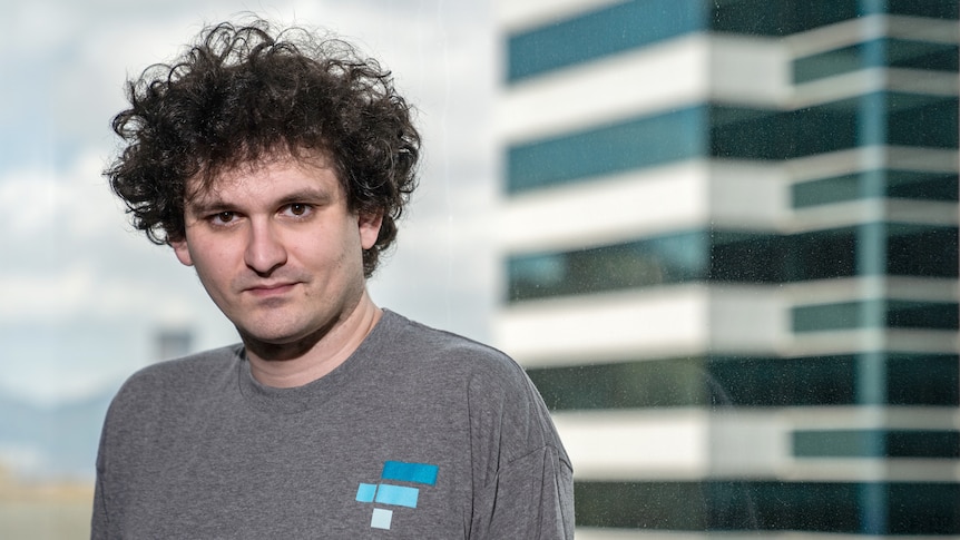 A man with big curly dark hair stands in front of a window overlooking buildings