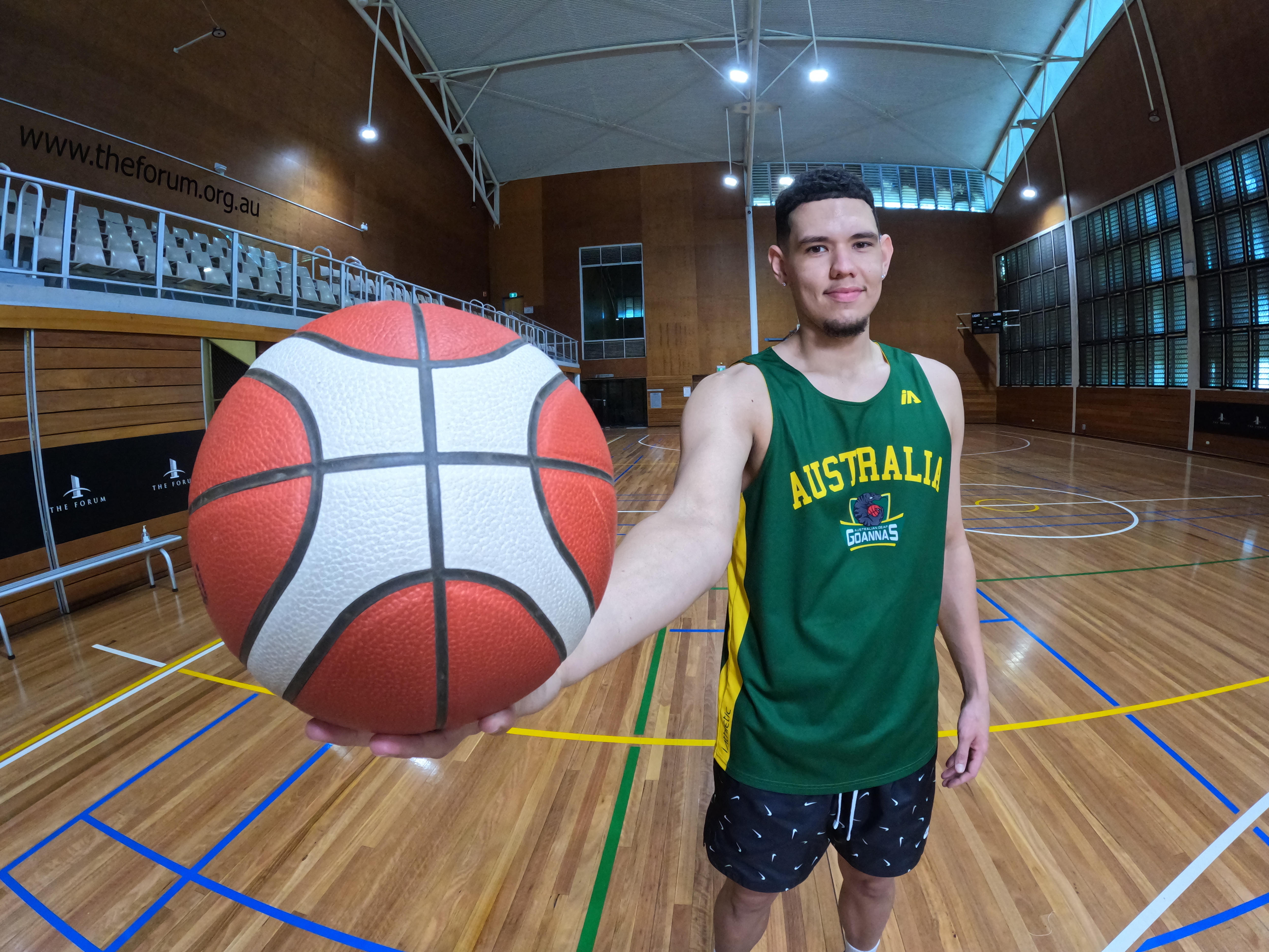 Jarrod is wearing a green "Australia" basketball jersey, and smiling while he holds a ball up close to the camera.
