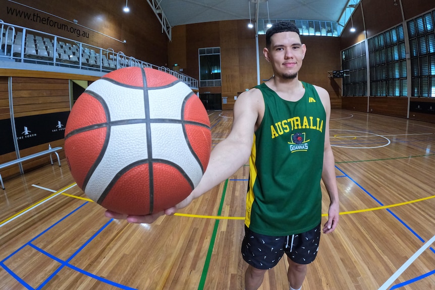 Jarrod is wearing a green "Australia" basketball jersey, and smiling while he holds a ball up close to the camera.