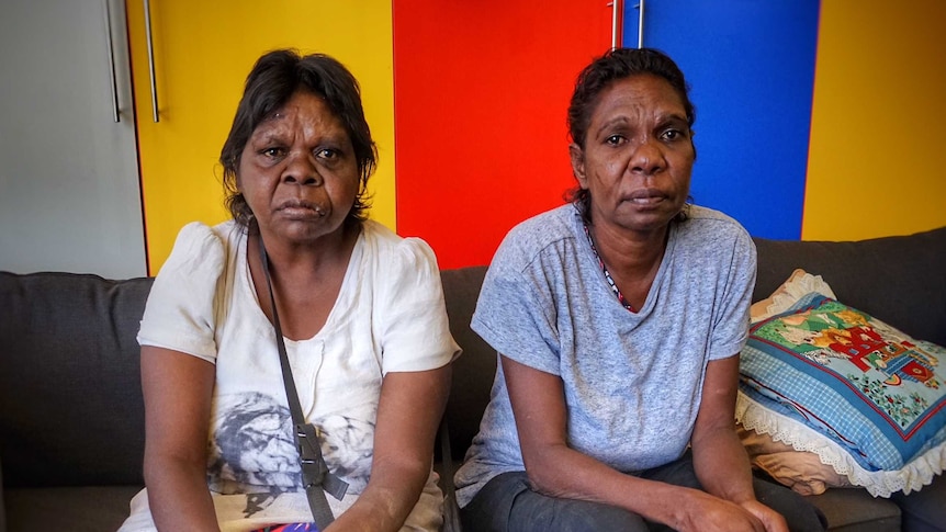 The two woman sit on a couch and look at the camera