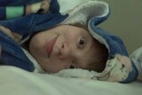 A woman wearing a blue dressing gown with the hood up lies on her side, looking at the camera, on a bed