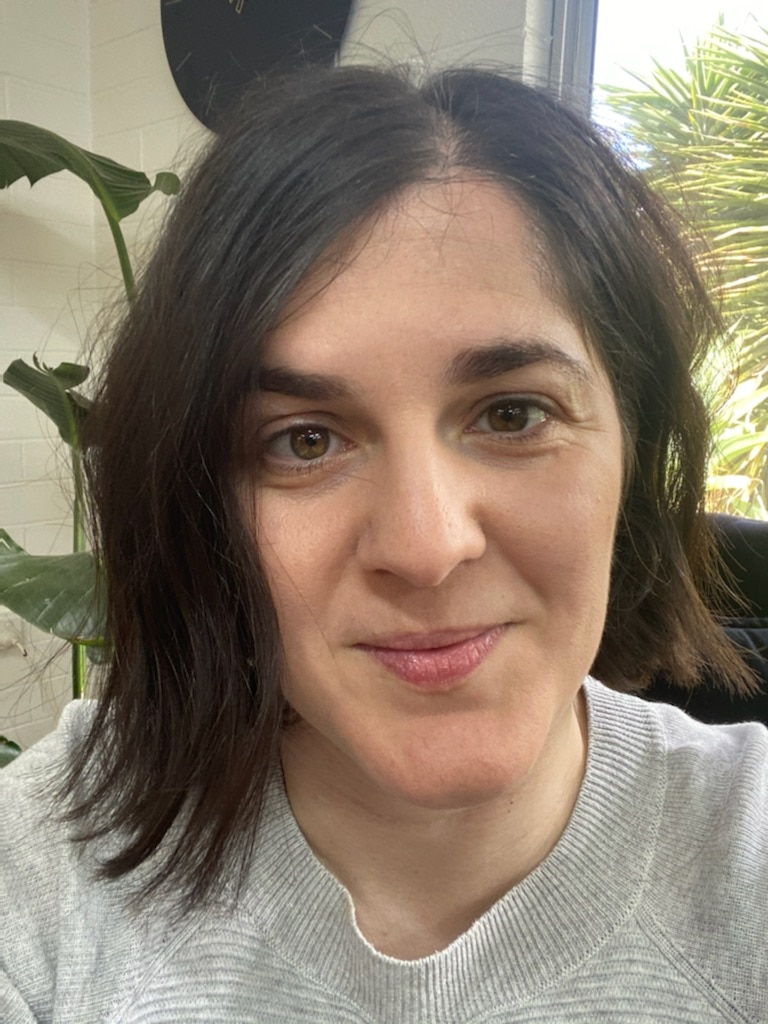 Dark haired woman smiling with an indoor plant and window in the background.