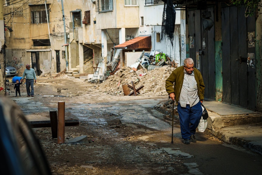 A man walks down the street marked by rubble with a walking stick.