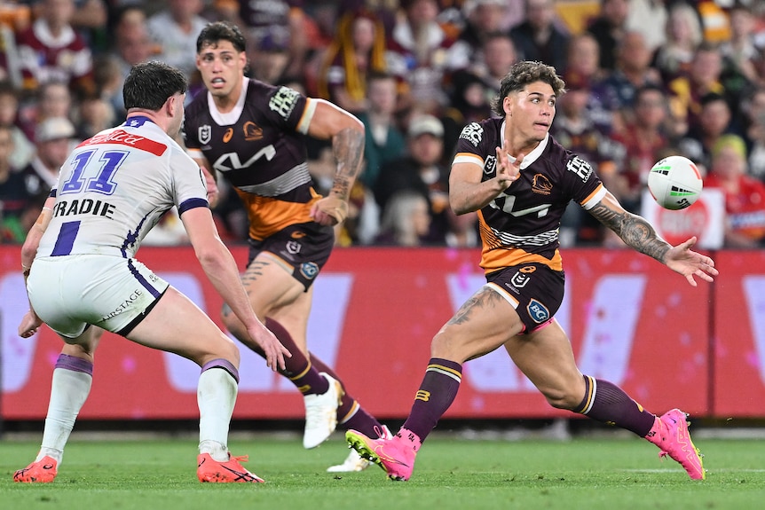 A Brisbane Broncos NRL player looks to his left as he sends a pass away as a defender braces to tackle him.