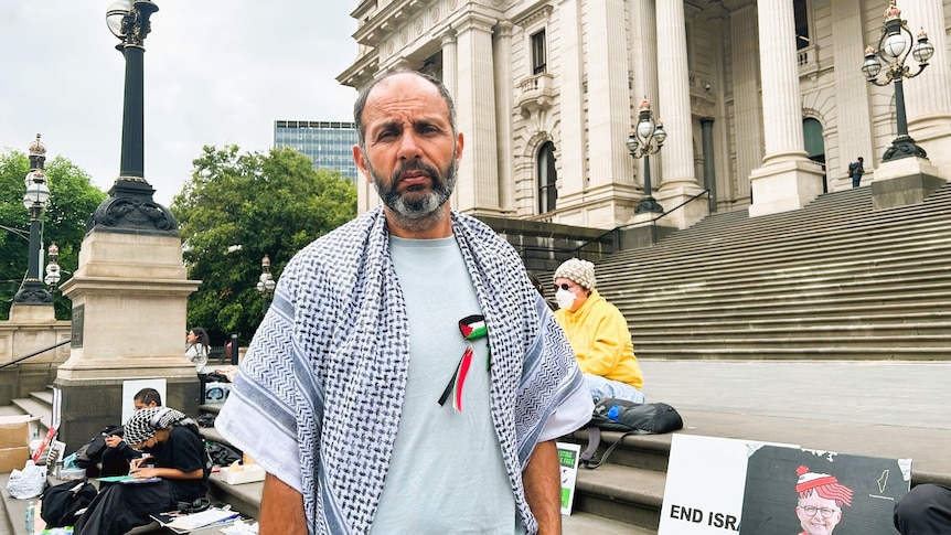 A man stands wearing a palestinian scarf, therer are signs behind him and parliament house is shown in the background.