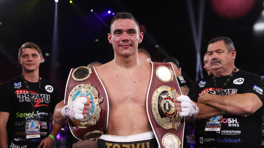 Australian boxer Tim Tszyu smiles as he holds two boxing title belts, one over each shoulder.  