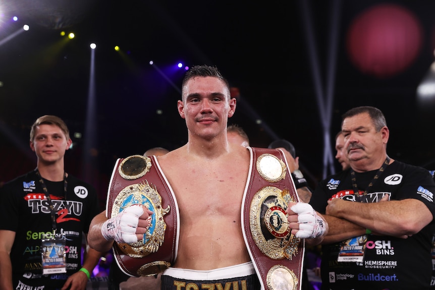 Australian boxer Tim Tszyu smiles as he holds two boxing title belts, one over each shoulder.  