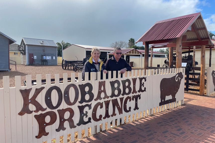 Woman and man lean over picket fence with large sign reading Koobabbie precinct