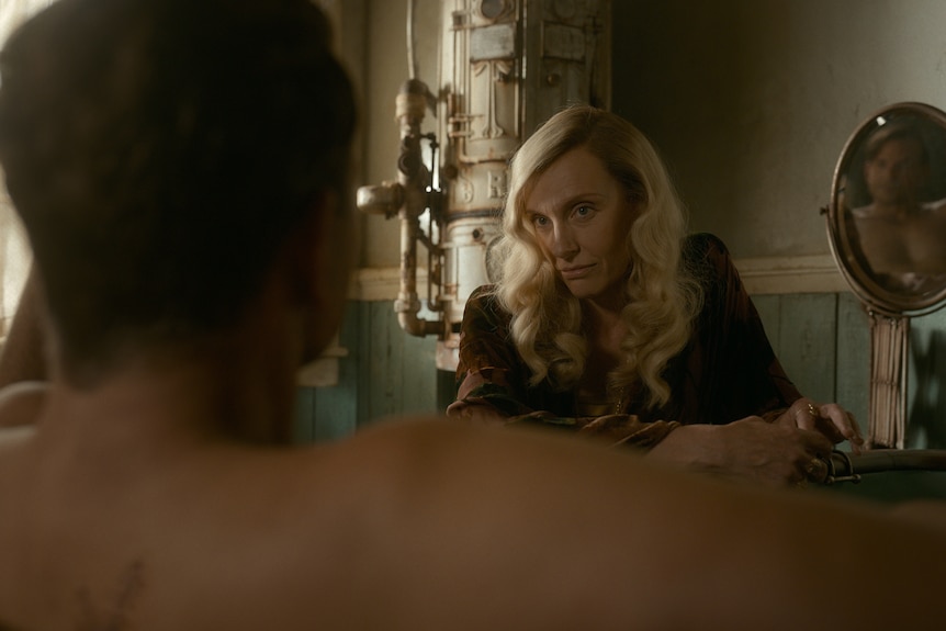 A 40-something blonde woman in 30s garb looks seriously into the face of a man who is in the bath, his back to the camera