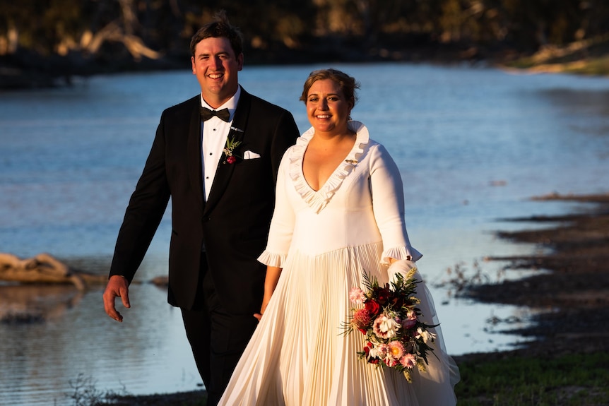 A bride and groom smile at camera as they walk past a river