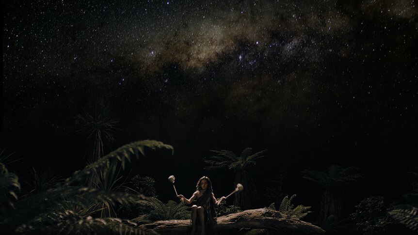 Under a starry night sky a young boy surrounded by ferns sits on fallen tree trunk swings small tethered weights in each hand.
