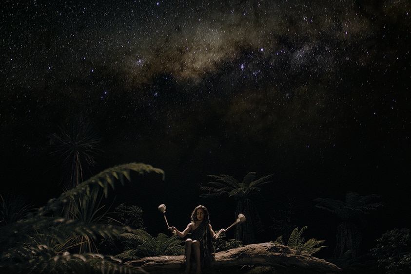 Under a starry night sky a young boy surrounded by ferns sits on fallen tree trunk swings small tethered weights in each hand.