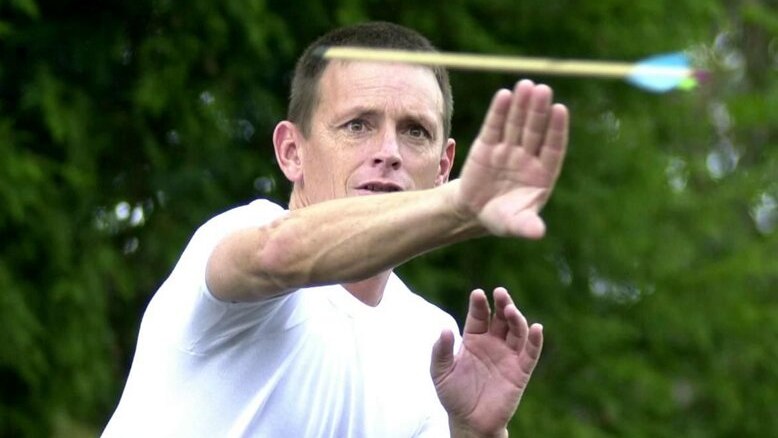 A man in a white shirt focuses on an arrow, holding his hand out in front to catch it