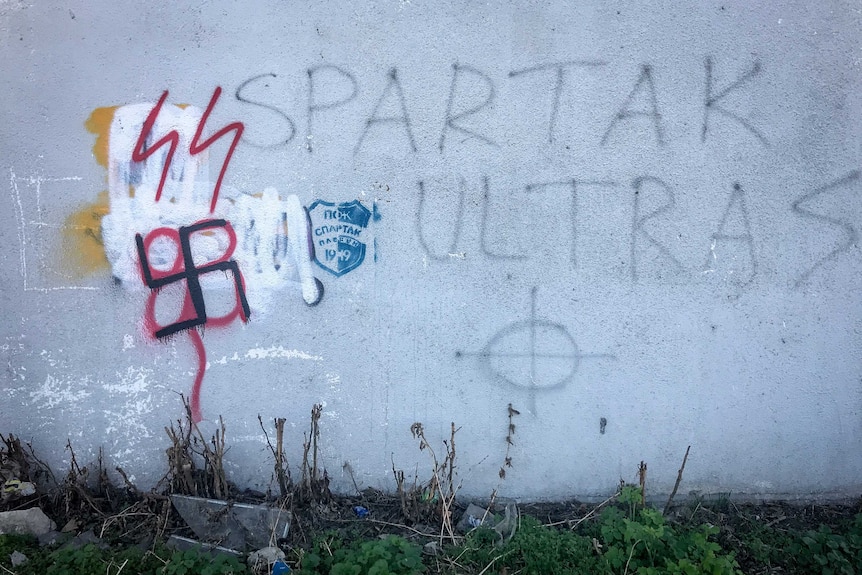 A black swastika is spray painted onto grey wall with two lightning bolts above. Weeds grow on the ground below.