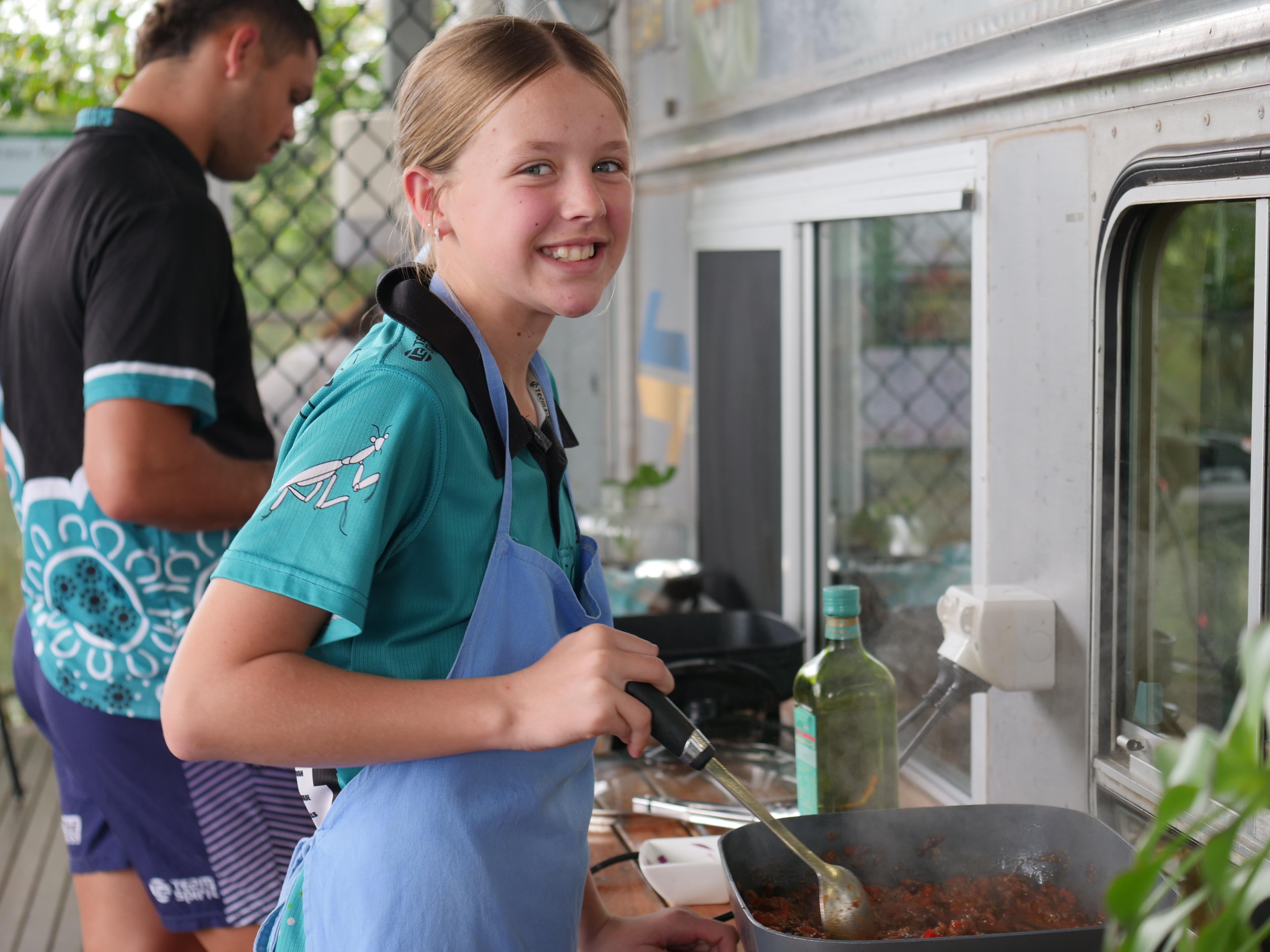 A primary school girl stands on a deck cooking food in a pan, wearing an apron and smiling.
