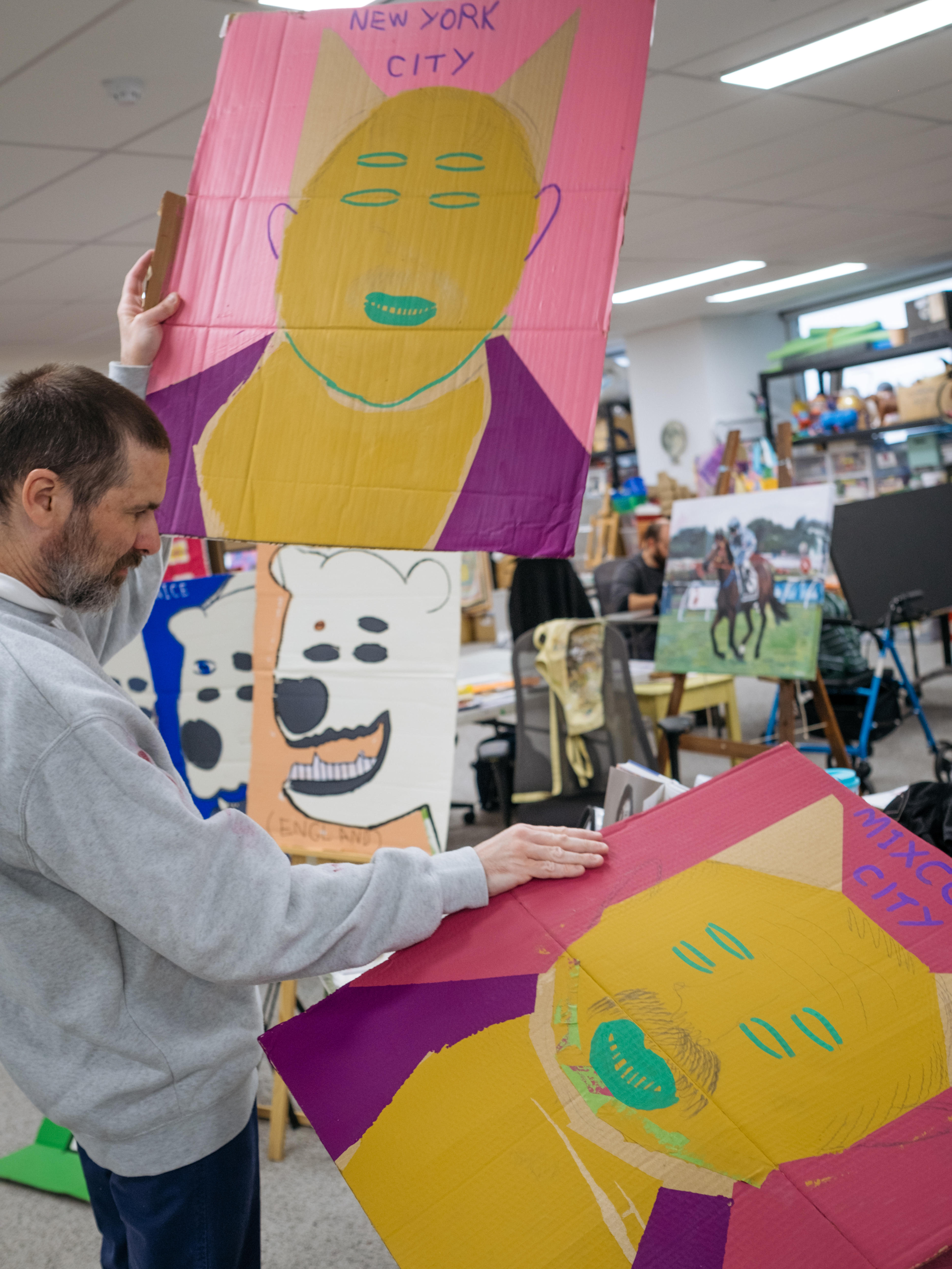 The artist in the studio holds two artworks at different angles. Painted on cardboard, they are brightly coloured and abstract