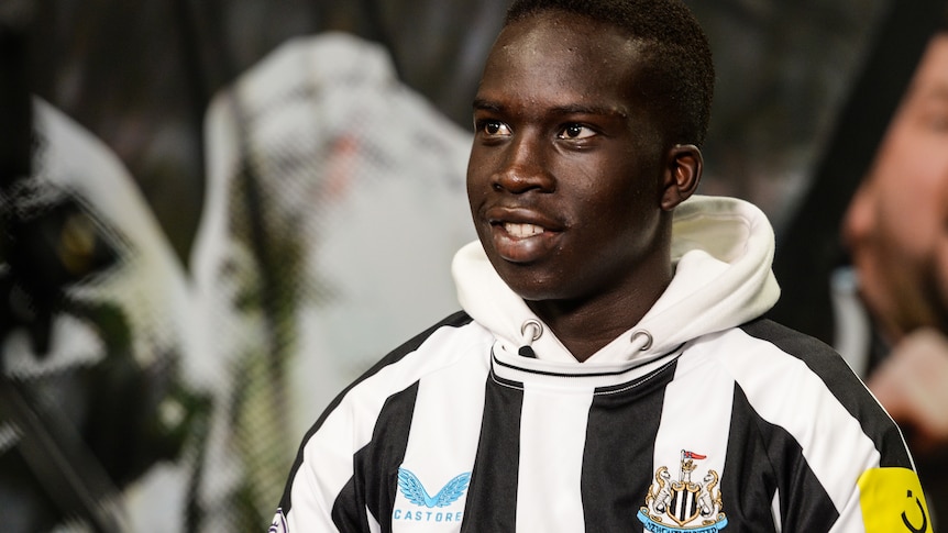 Garang Kuol smiles and looks upwards to his right in a Newcastle United shirt and hoodie