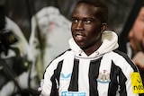 Garang Kuol smiles and looks upwards to his right in a Newcastle United shirt and hoodie