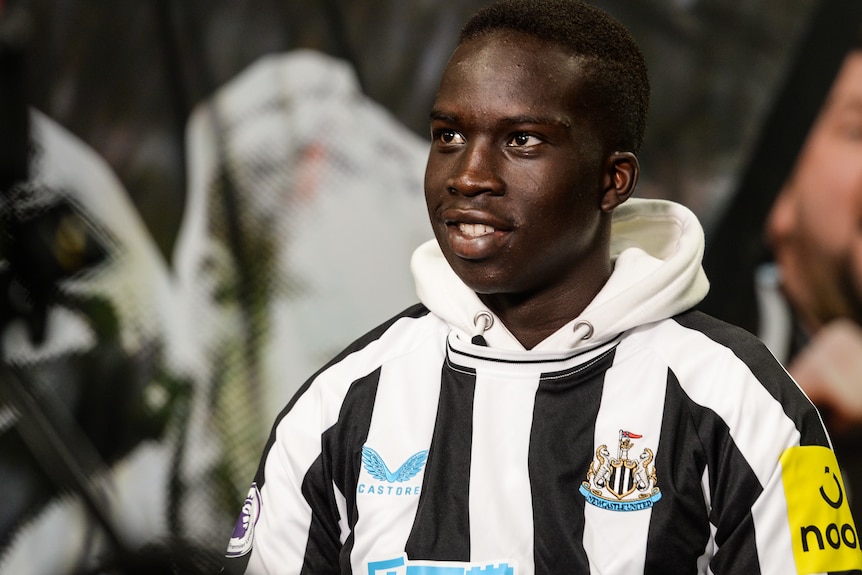 Garang Kuol smiles and looks upwards to his right in a Newcastle United shirt and hoodie