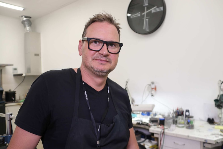 A mid shot of jeweller Rohan Milne standing in his workshop posing for a photo wearing a black shirt, black apron andspectacles.