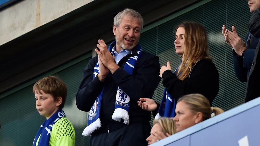 Roman Abramovich in a Chelsea club scarf claps, while standing next to a blonde teen girl 