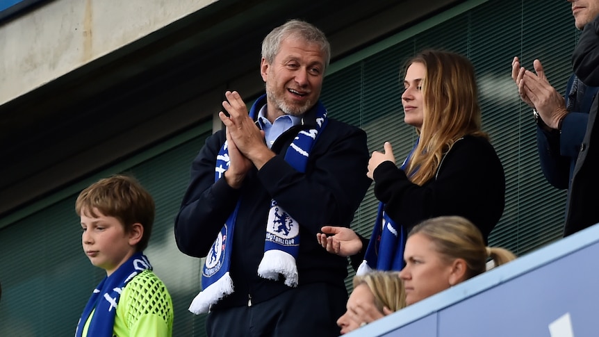 Roman Abramovich in a Chelsea club scarf claps, while standing next to a blonde teen girl 