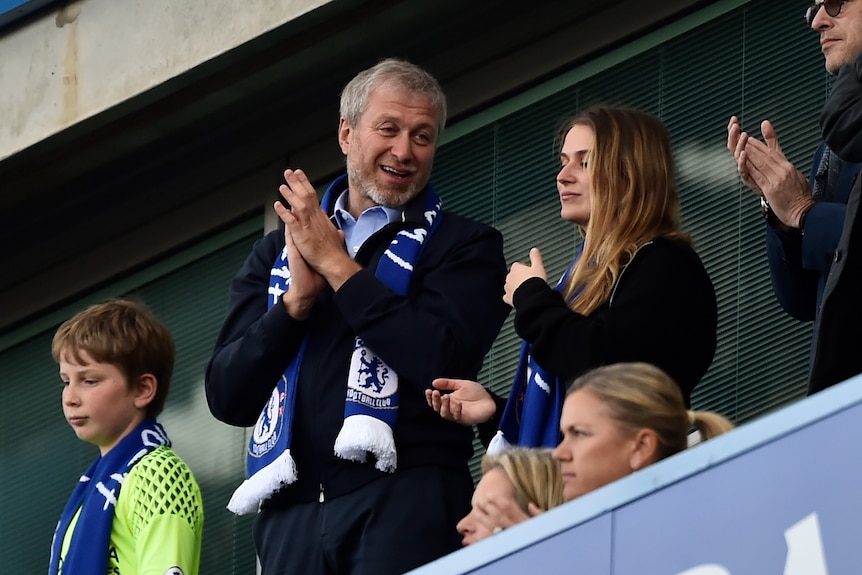 Roman Abramovich in a Chelsea club scarf claps, while standing next to a blonde teen girl 