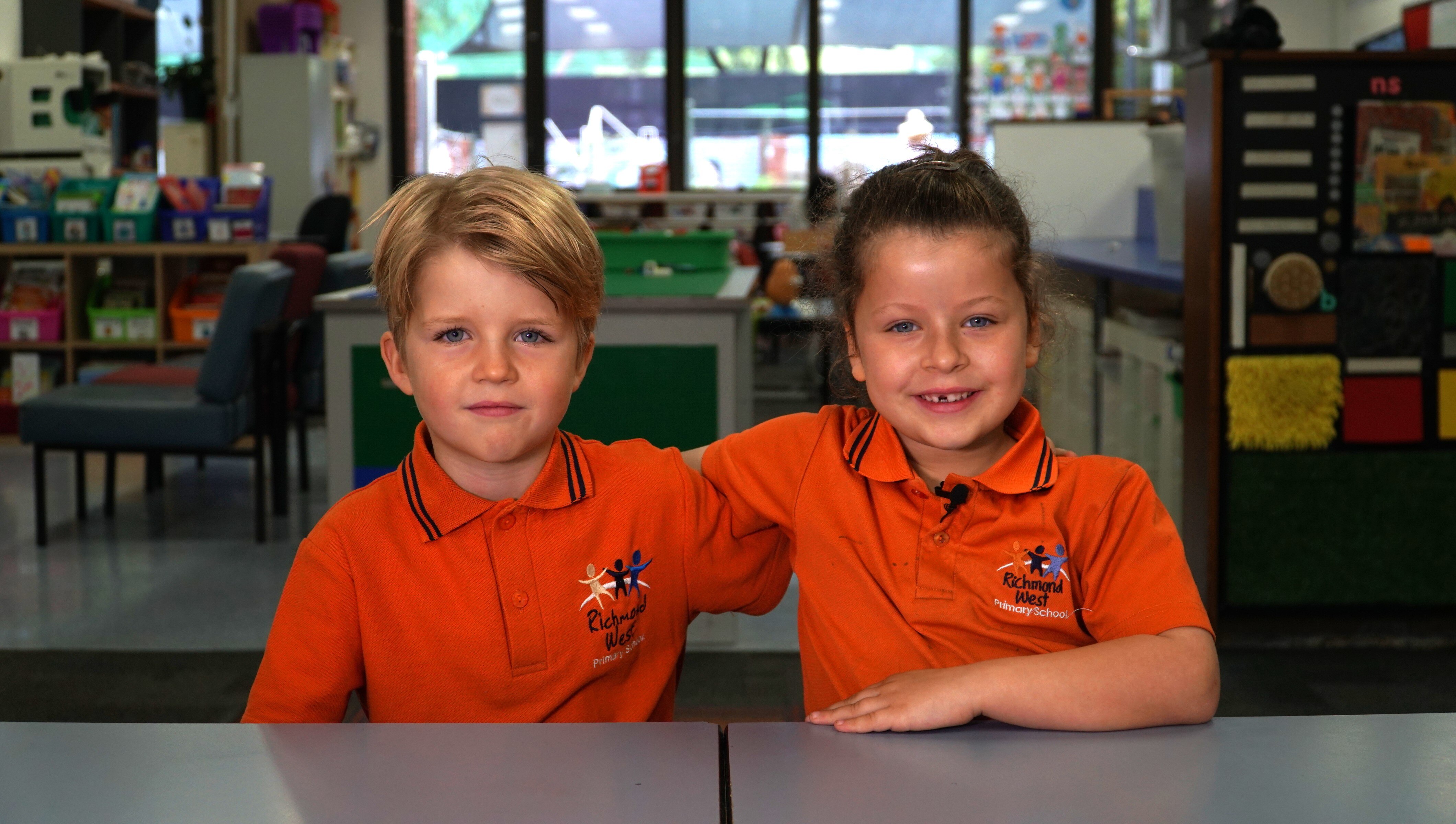 Two young kids, arm in arm, sit in a classroom.