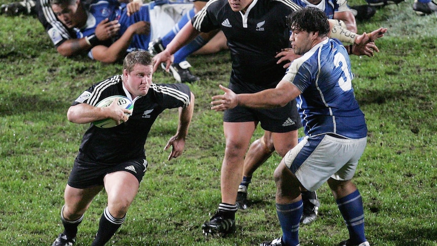 New Zealand rugby union player tucks the ball under his arm and runs around a Samoan defender during a game.