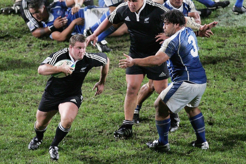 New Zealand rugby union player tucks the ball under his arm and runs around a Samoan defender during a game.