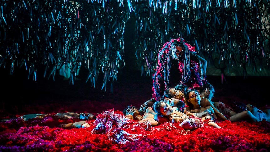 Scissors dangle above a group of dancers lying on the ground enveloped in red feathers.