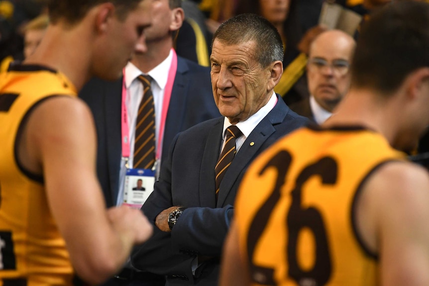A man in a suit stands with his arms folded among Hawthorn players.