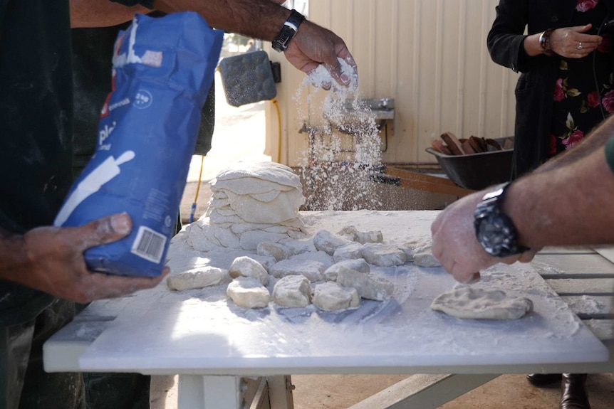 A man sprinkles flour over dough while another kneads.