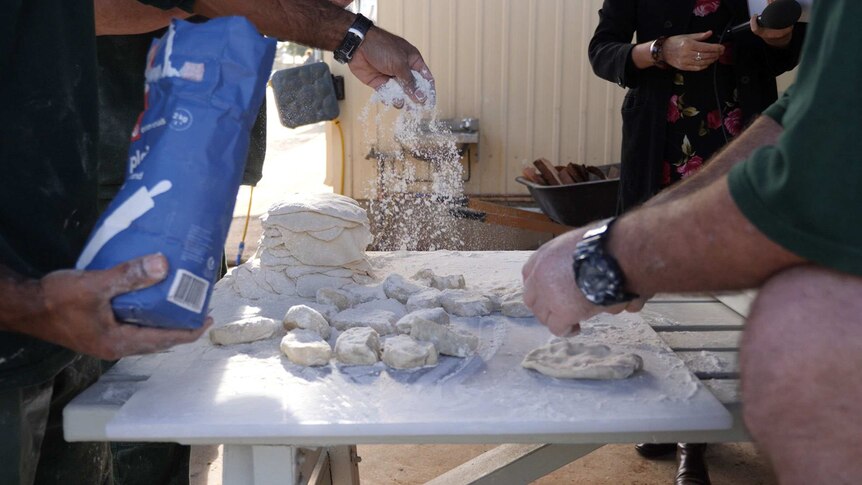 A man sprinkles flour over dough while another kneads.