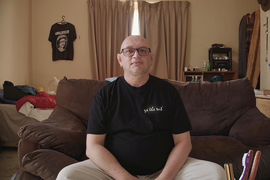 A bald man wearing glasses sits on a couch and looks at the camera