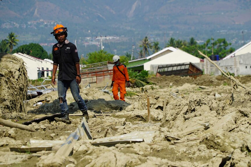 Two male rescue workers walk across a field of uneven mud, with a house half buried, on an angle in the background