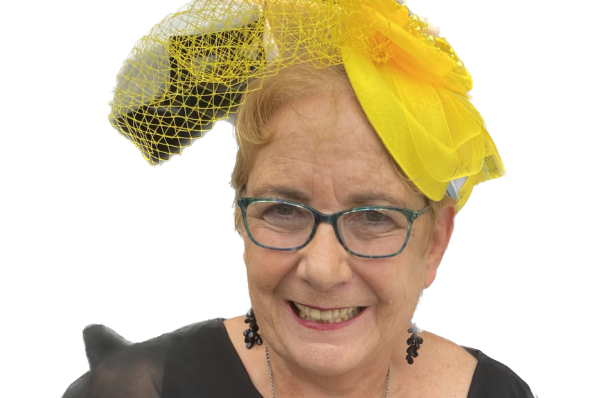 A headshot of Maxine, who is smiling and wearing glasses and a yellow fascinator on her head.