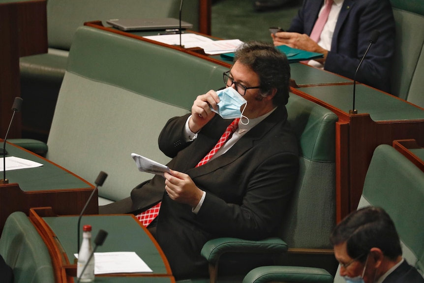 George Christensen sits among the green benches of the Lower House. He holds a blue surgical mask over his mouth and nose.