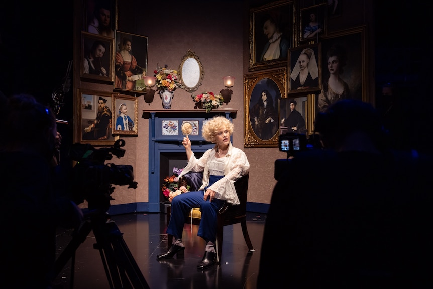 Woman dressed as a young man with curly flaxen hair sits on chair and holds mirror surrounded by portrait paintings and cameras