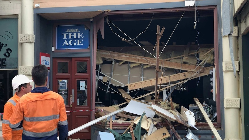 Men in hi-vis appraise the front of a newsagency that has been badly damaged.