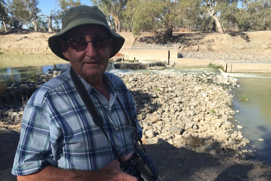 A man with a hat and glasses holds a camera. Behind him is a shallow Darling River