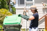 A woman wearing a large-brim hat and gardening gloves places old branches in a green waste recycling bin.