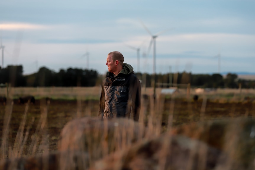 Man in jacket stands walks in early morning light with farm behind him