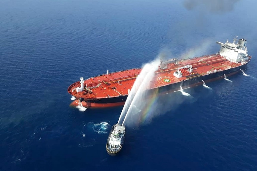 A boat sprays two large streams of water at a stricken oil tanker that is on fire in open water.